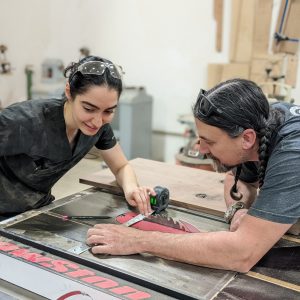a man in a gray shirt leans over a table saw, helping a woman in a black shirt holding a measuring tape