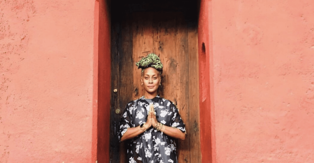 picture of a yoga teacher inside of a red doorway with wood behind her and her hands together in front of her heart looking at the camera